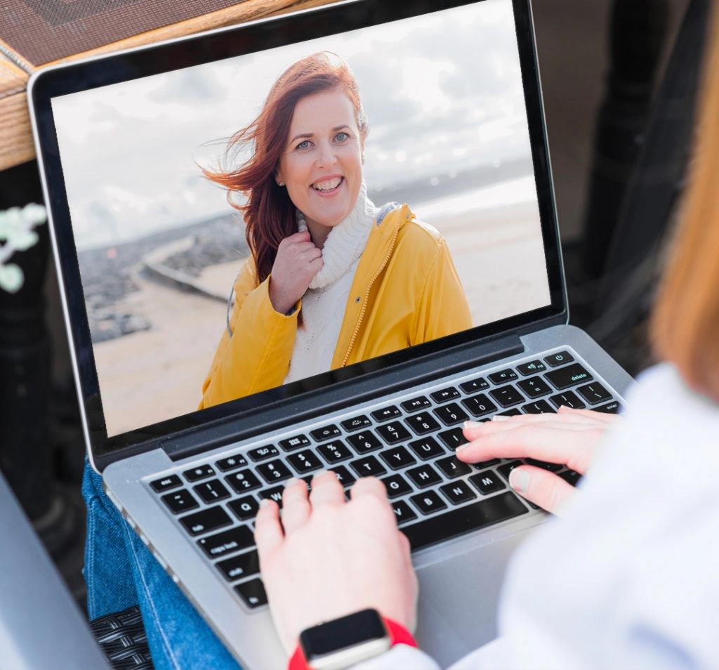 Person video chatting on a laptop with a woman in a yellow jacket by the beach, smiling at the camera.