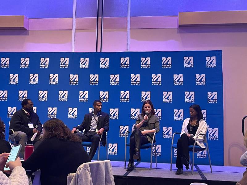 Panel discussion at UMass Lowell event with four speakers on stage, engaging in conversation before a seated audience.