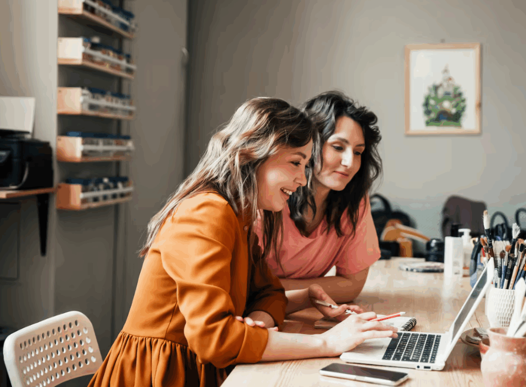 Two women smiling while working on a laptop at a wooden desk in a cosy home office.